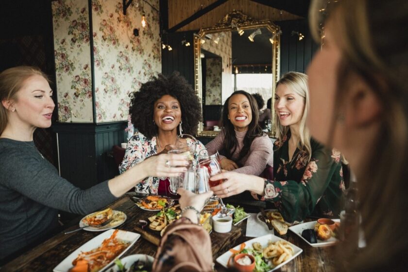 Three women at a sitting at a beautifully set table full of food clinking their glasses together 