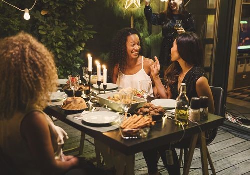 Three women sit outside at a candle lit table at a dinner party laughing